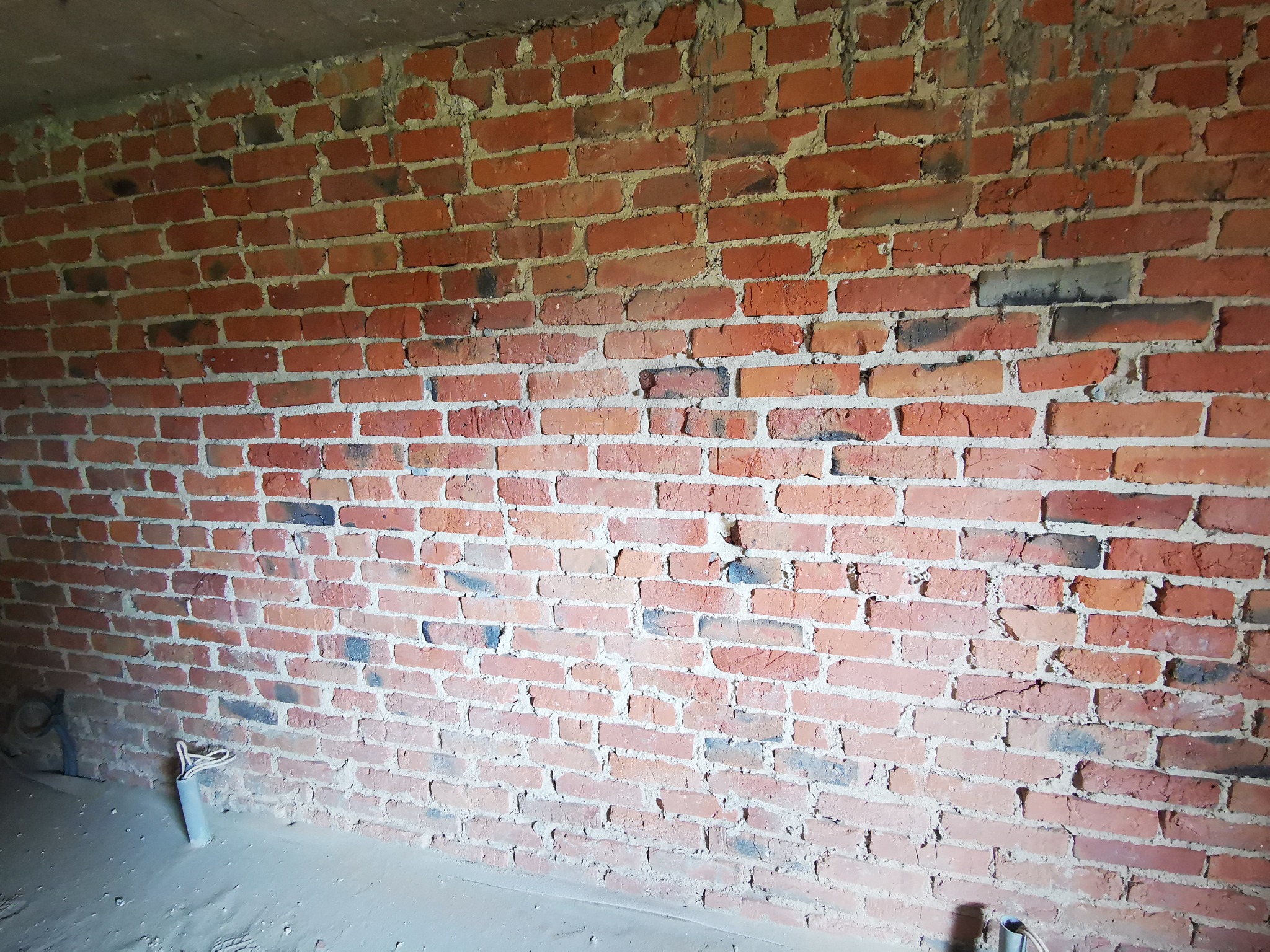 Surowa ściana z czerwonej cegły z widocznymi przewodami elektrycznymi wystającymi z podłogi, ukazująca stan przed malowaniem lub wykończeniem.