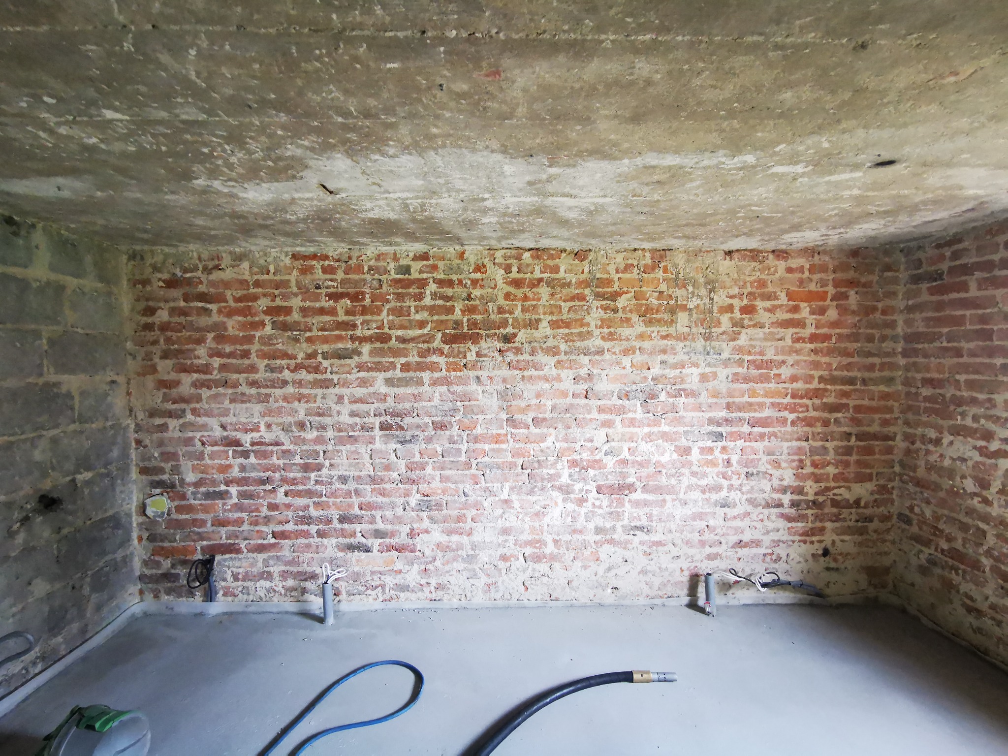 Surowe wnętrze pokoju z ceglaną ścianą, surowym betonowym sufitem i świeżo wylaną podłogą. Widoczne wystające instalacje i węże na podłodze.