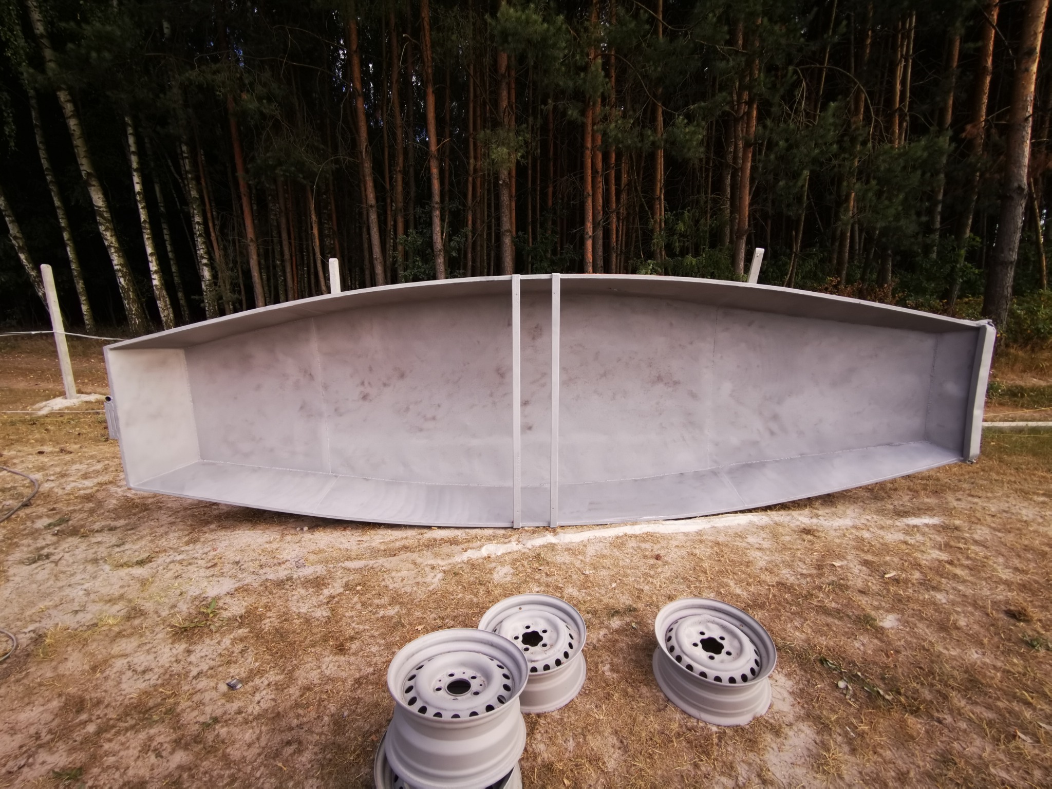 Metalowa konstrukcja w kształcie łodzi, odwrócona dnem do góry, po obróbce, leżąca na trawie obok trzech metalowych felg samochodowych pomalowanych na szaro, na tle lasu.