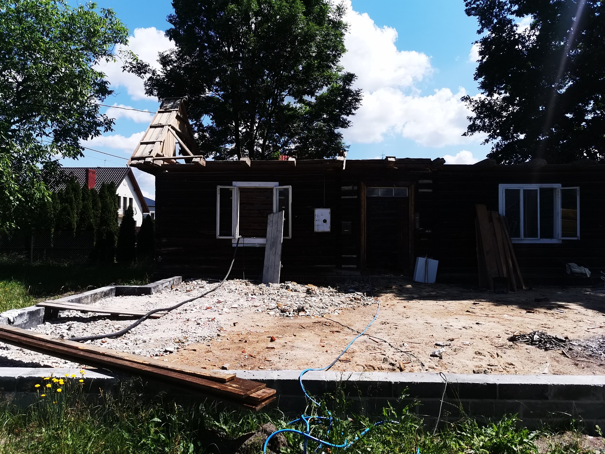 Drewniany dom w trakcie remontu, z odsłoniętą konstrukcją dachu i fundamentami, otoczony gruzem i zielenią.