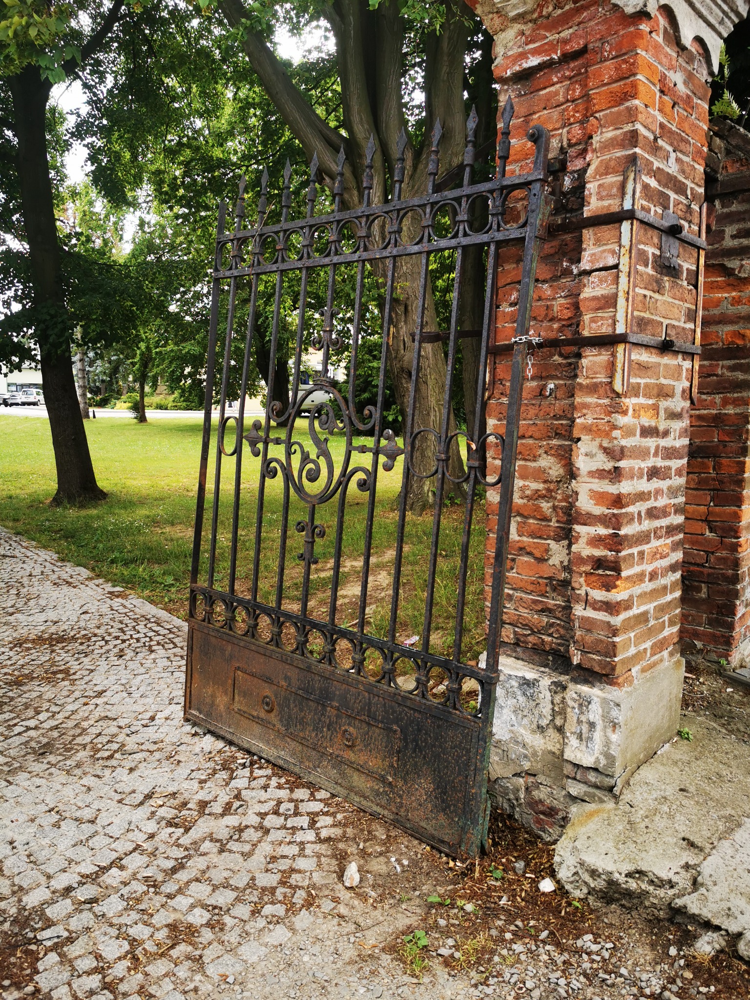 Otwarta, zardzewiała, żeliwna brama z ozdobnymi elementami, oparta o ceglany filar, na tle zielonego trawnika i drzew.