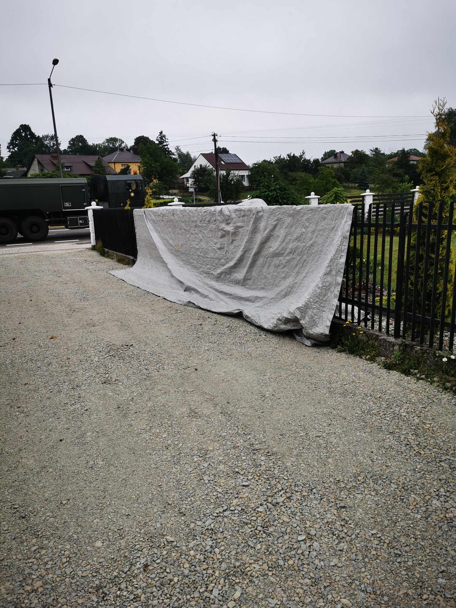 Czarna metalowa brama ogrodzeniowa zabezpieczona szarą plandeką ochronną, na żwirowym podłożu, z widocznym pojazdem wojskowym w tle.