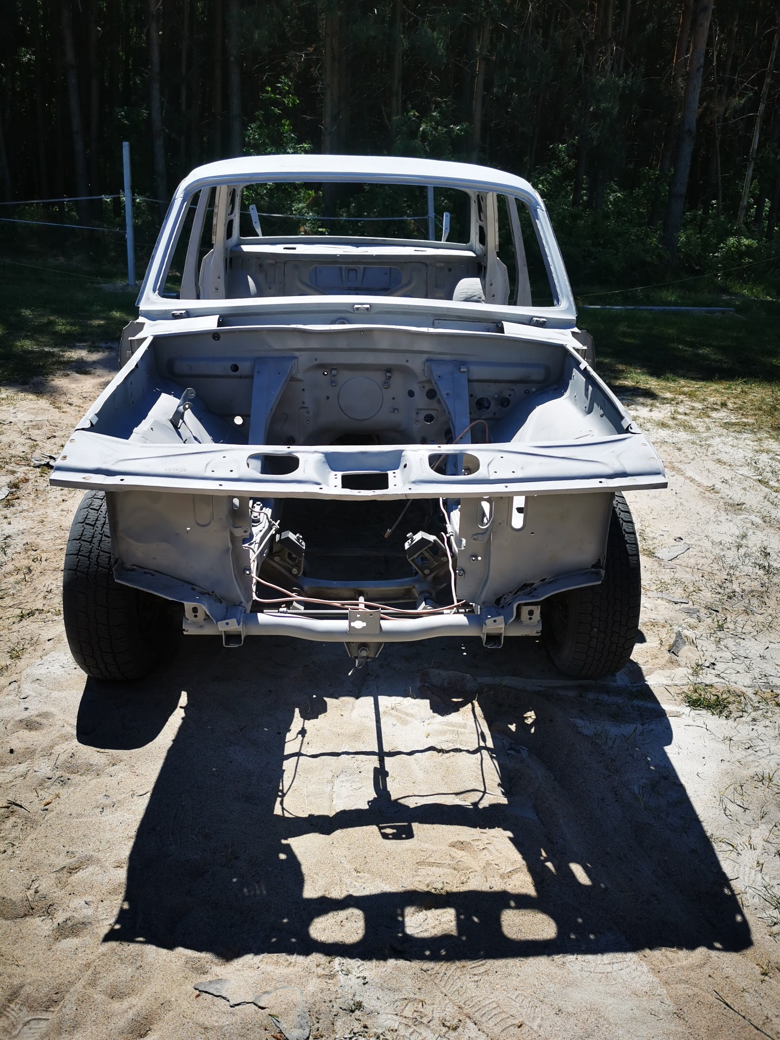 Przód karoserii samochodu po piaskowaniu, z widocznymi elementami zawieszenia i fragmentami opon, na piaszczystym podłożu w słoneczny dzień.