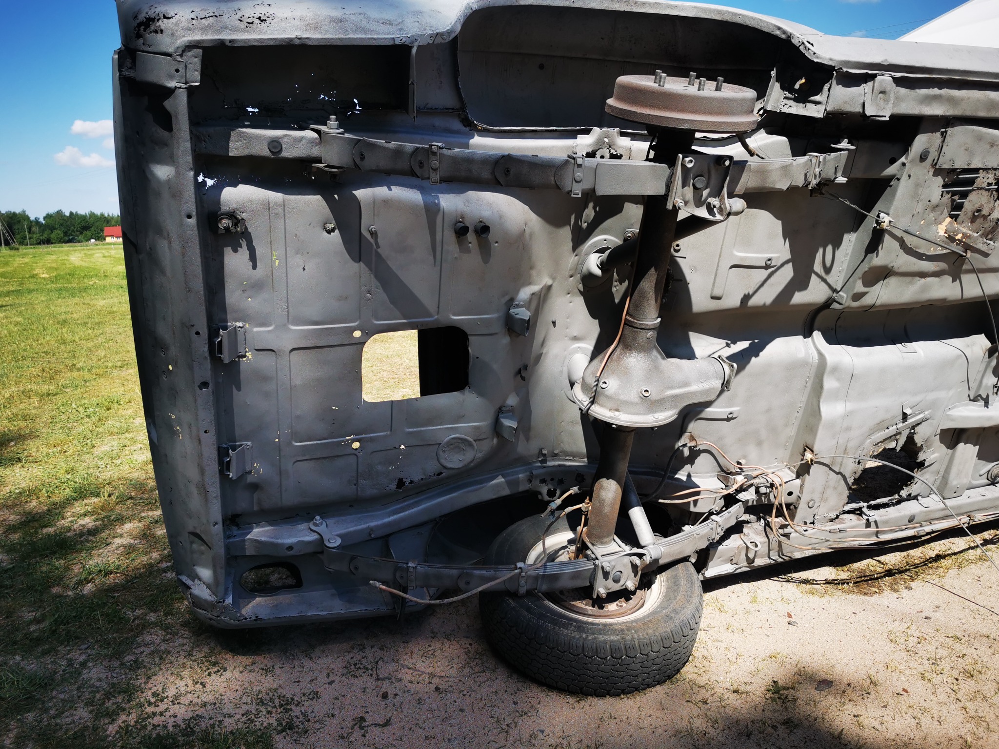 Podwozie samochodu osobowego odwrócone do góry kołami, po wstępnym oczyszczeniu z rdzy, widoczne ślady korozji i ubytki, na tle trawy i błękitnego nieba.