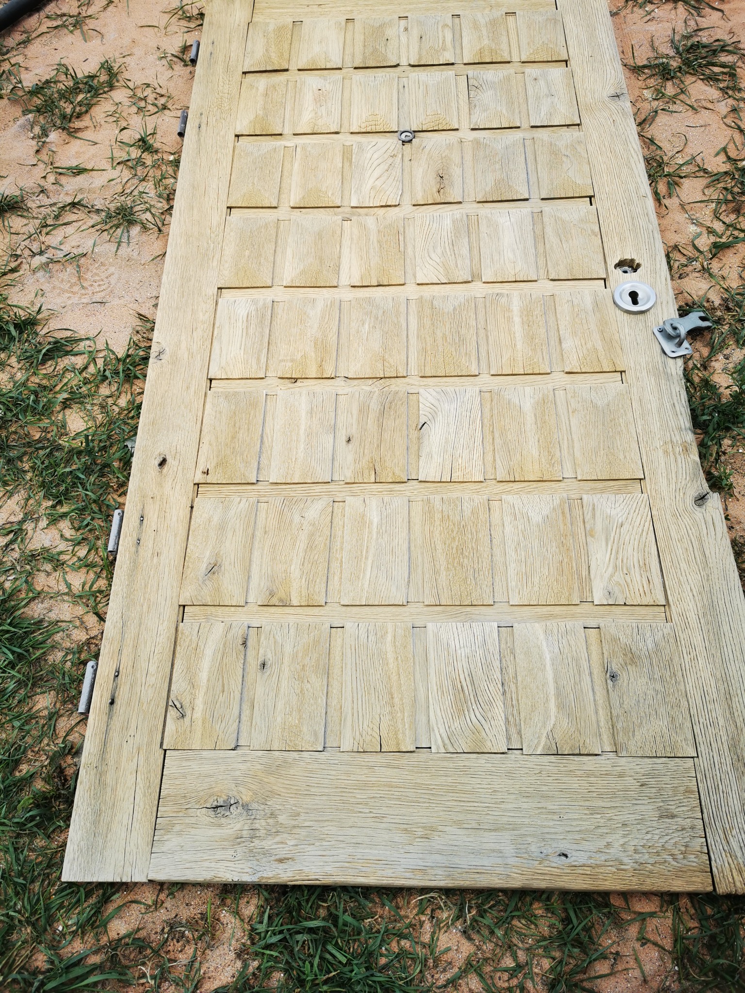 Drewniane drzwi z panelami, leżące na ziemi z piaskiem i trawą, widoczne ślady po usunięciu starej farby lub lakieru, przygotowane do dalszej obróbki.