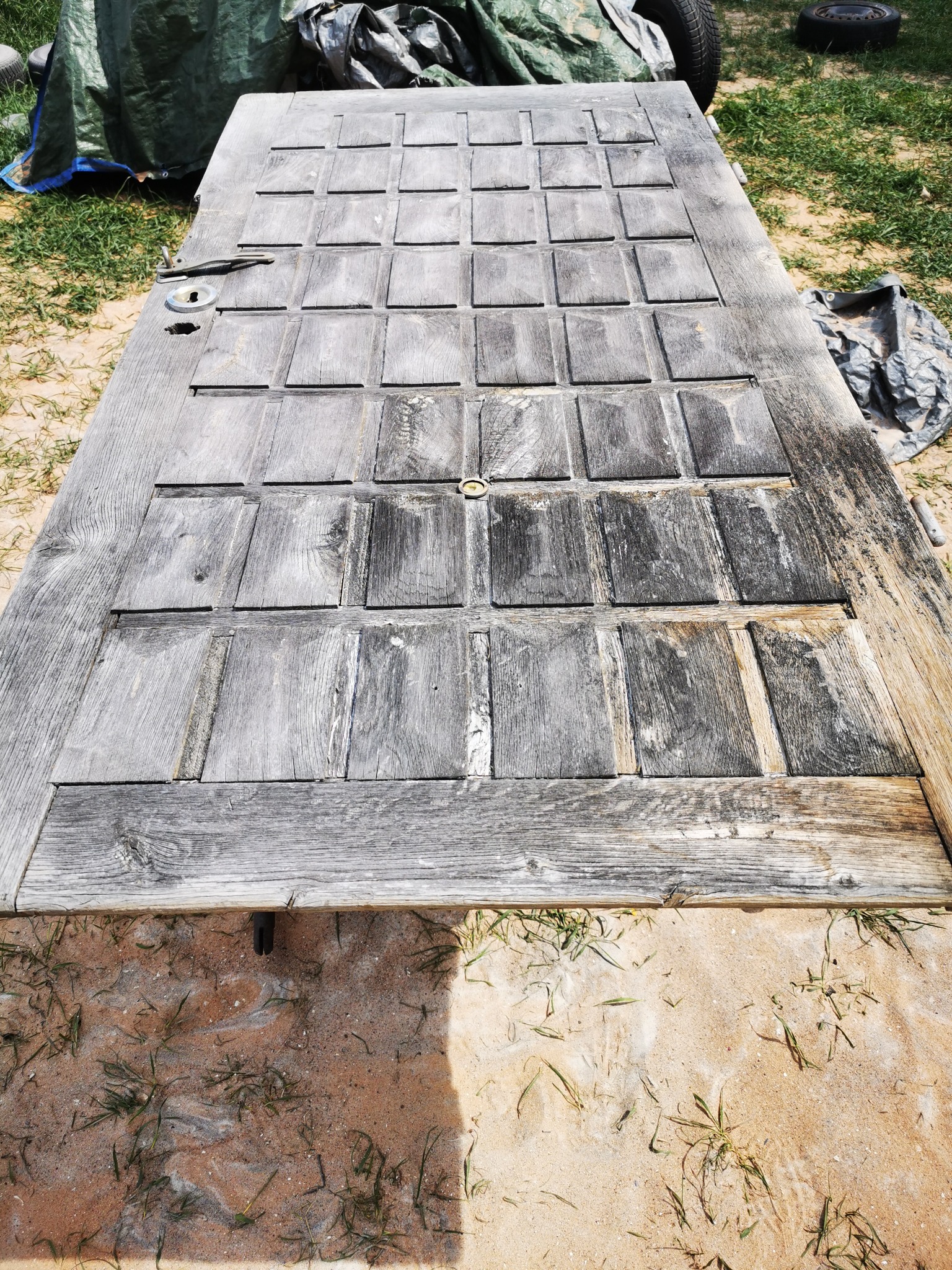 Stara, drewniana, panelowa drzwi leżąca na piasku, z widocznymi śladami zużycia i pozostałościami po okuciach.