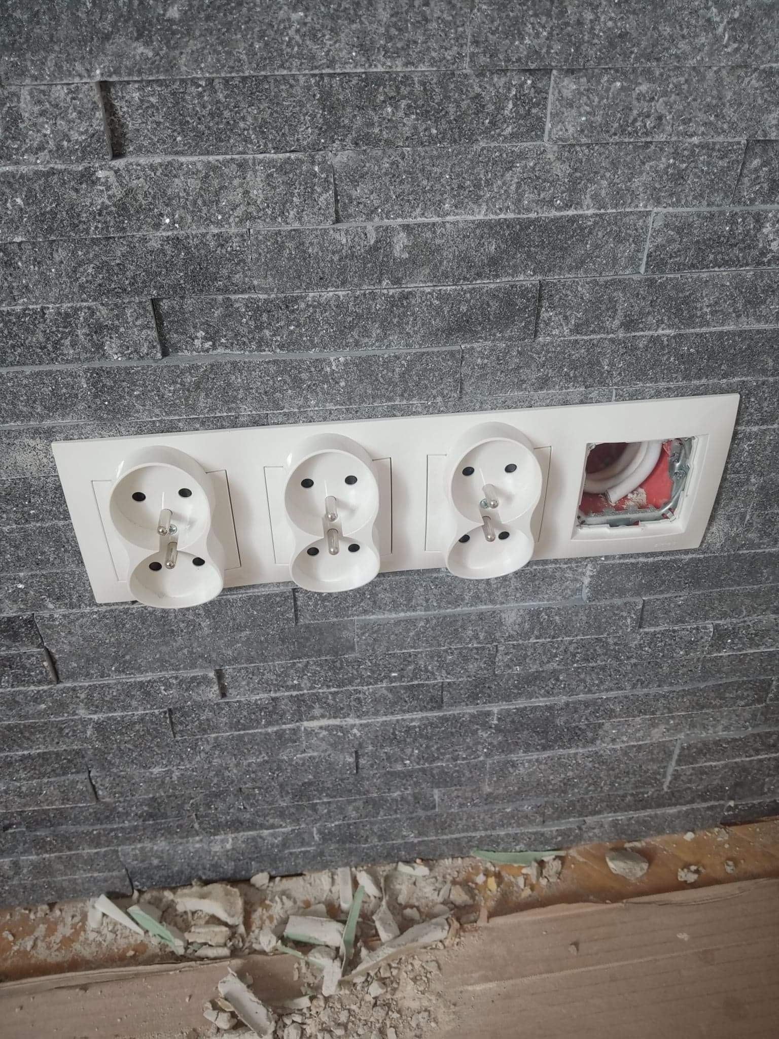 Montaż potrójnego gniazdka elektrycznego z bolcem na szarej, kamiennej ścianie, widoczne okablowanie w jednym z otworów, gruz na podłodze.