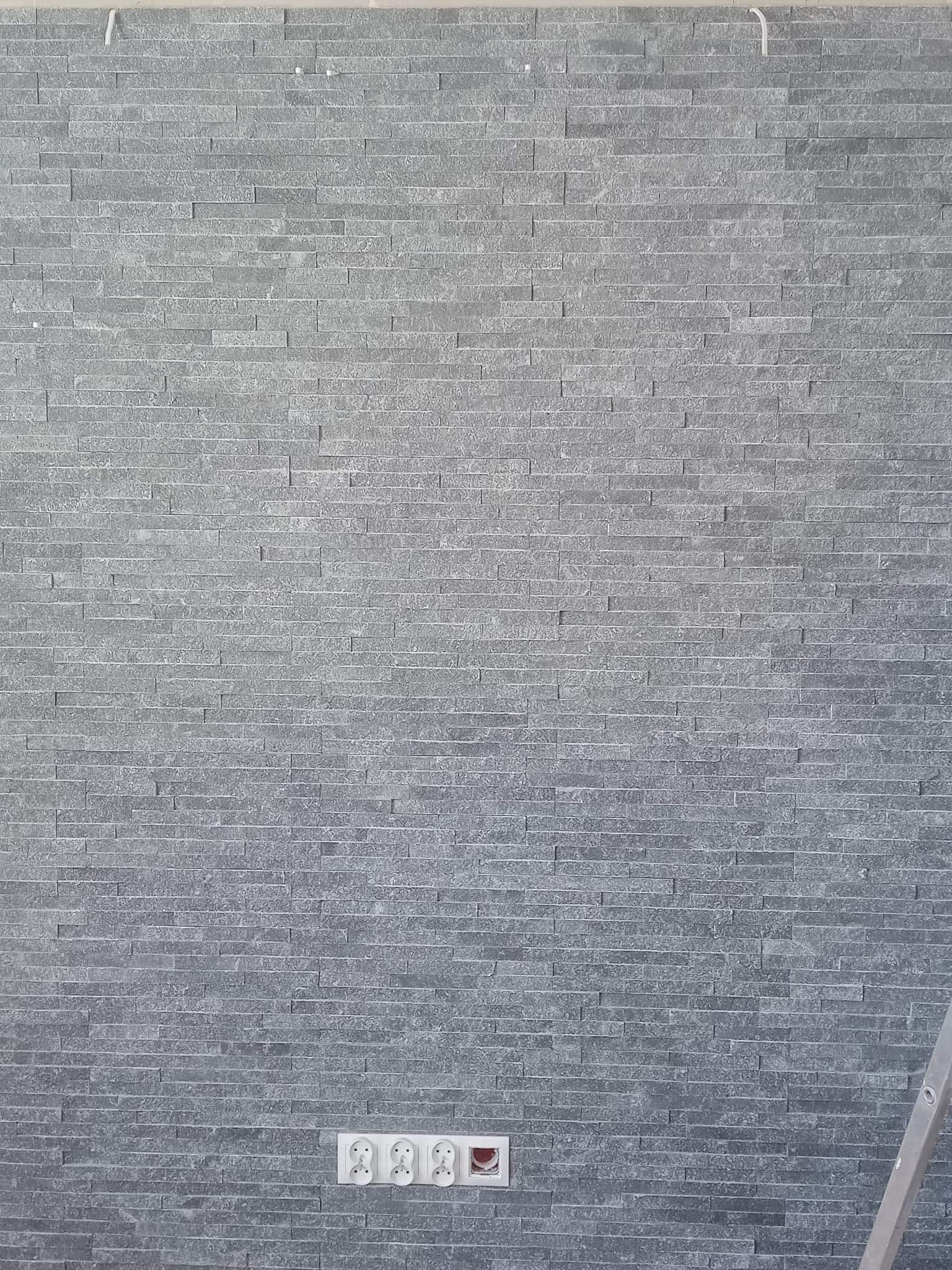 Ściana wykończona szarą okładziną kamienną o wąskich, podłużnych elementach, widoczne gniazdka elektryczne i fragment drabiny.