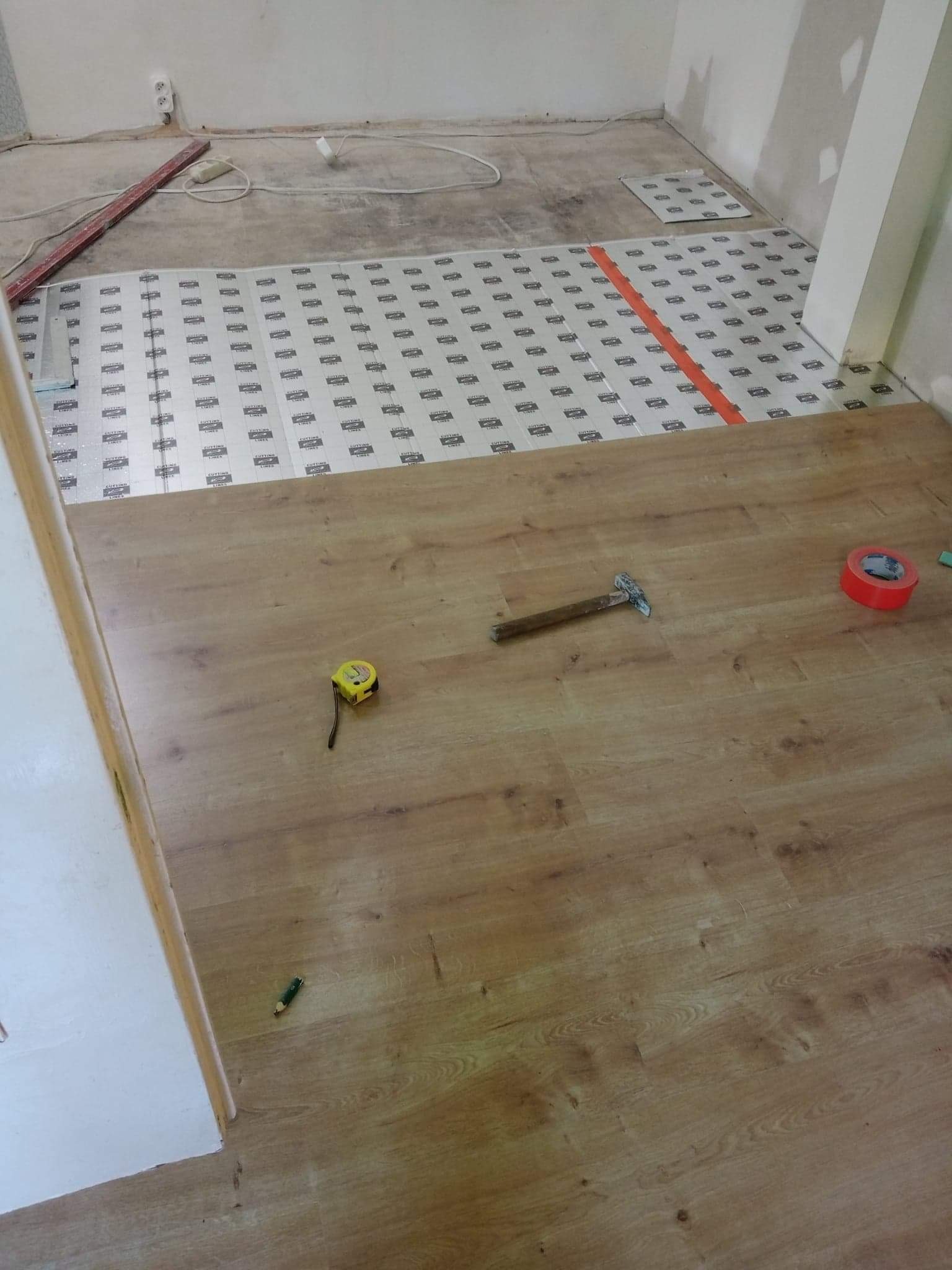 Układanie paneli podłogowych: widoczny podkład izolacyjny, częściowo ułożone panele w odcieniach brązu, młotek, miara zwijana i taśma klejąca na podłodze w trakcie prac wykończeniowych.