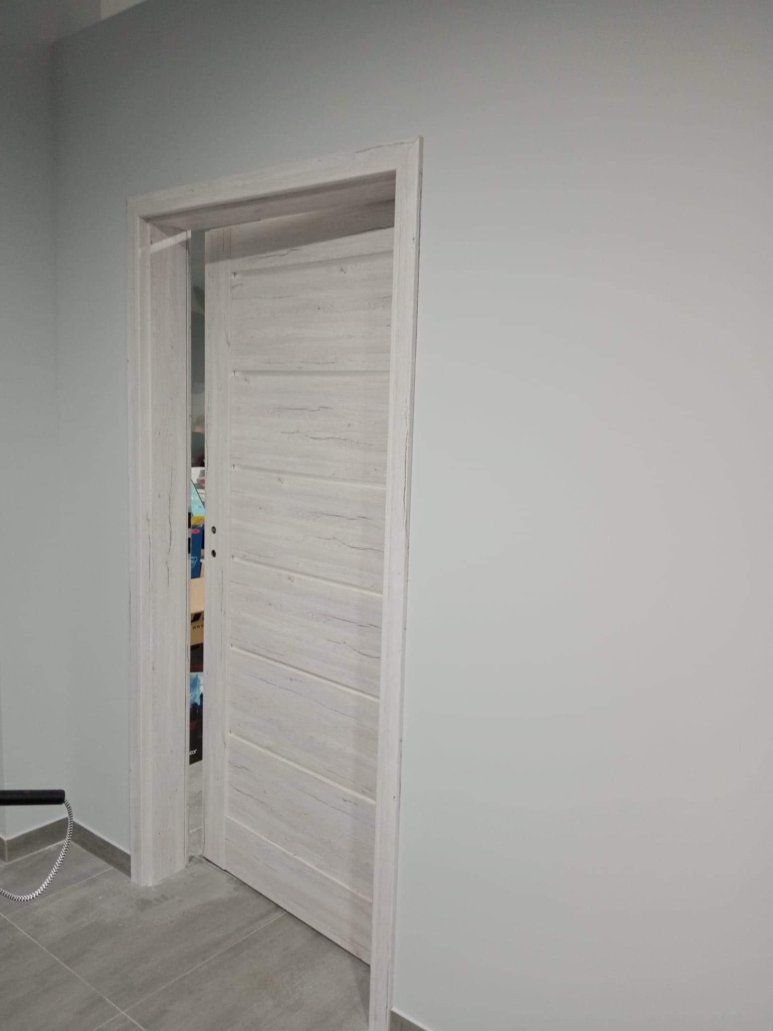 Nowe, białe, drewniane drzwi wewnętrzne z poziomymi rowkami, częściowo otwarte, widoczne szare płytki podłogowe i fragment ściany w tle.