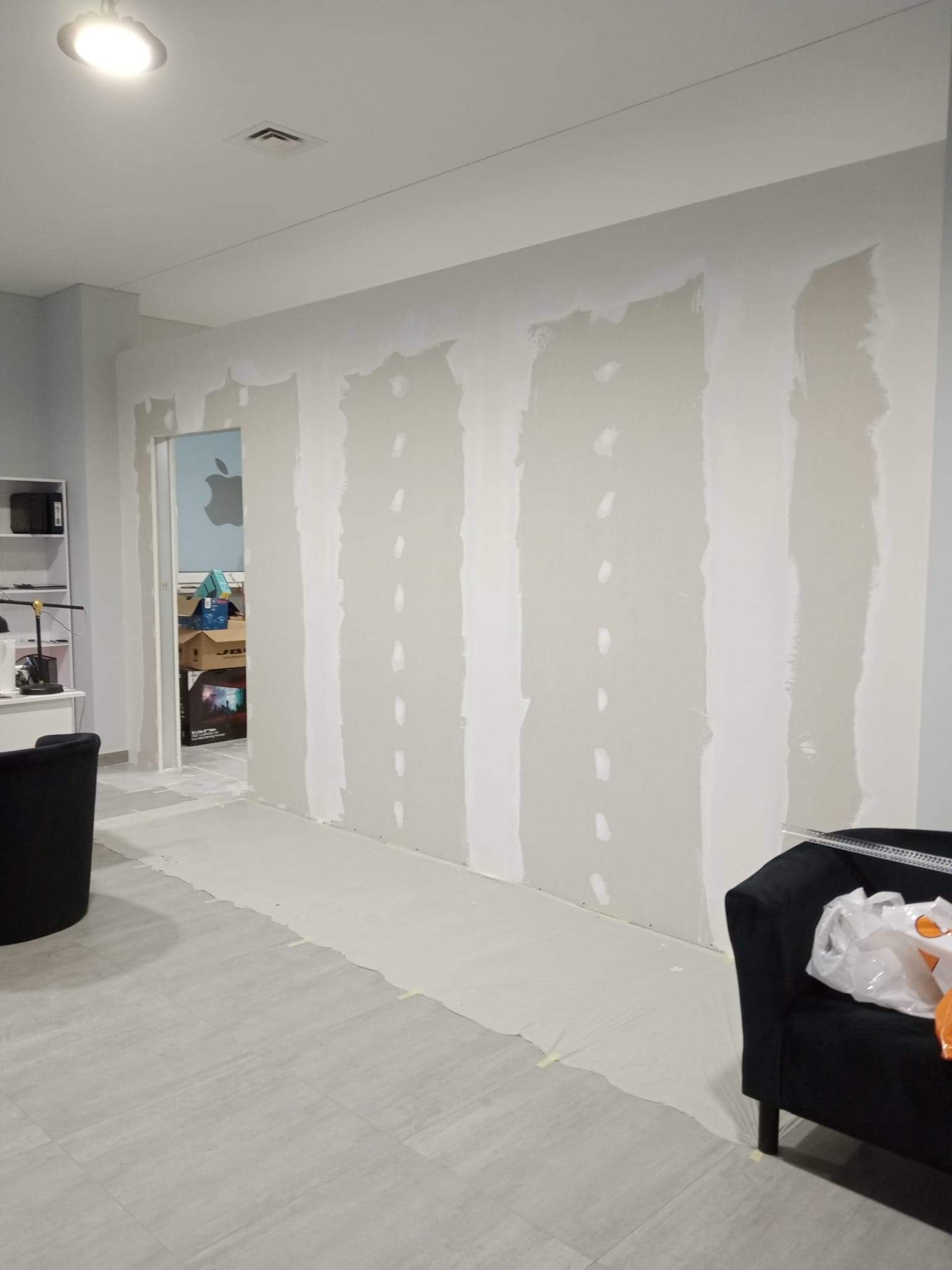 Przygotowana ściana do szpachlowania z widocznymi paskami gipsowymi, podłoga zabezpieczona folią malarską, w tle widoczne meble i sprzęt biurowy.