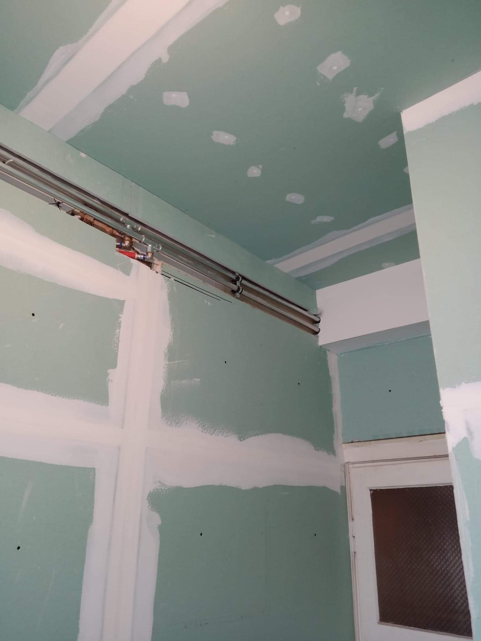 Wnętrze w trakcie prac wykończeniowych: widoczne płyty gipsowo-kartonowe, szpachlowane łączenia, instalacja hydrauliczna pod sufitem i fragment drzwi.