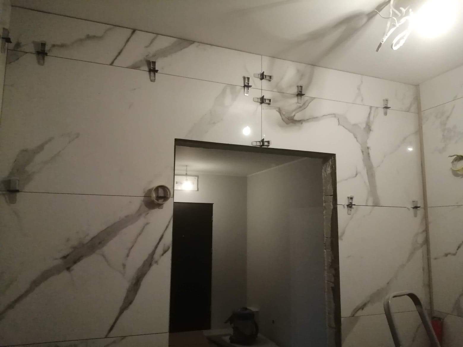 Płytki imitujące marmur na ścianie, z widocznymi klipsami poziomującymi i sznurkiem, obok otworu drzwiowego w trakcie prac wykończeniowych.