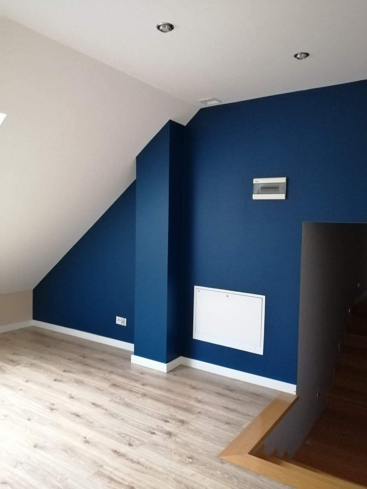 Poddasze po remoncie: ściana w kolorze głębokiego granatu, biała listwa przypodłogowa, jasna podłoga panelowa, widoczne elementy instalacji elektrycznej i skrzynka rozdzielcza.