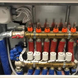 Rozdzielacz ogrzewania podłogowego z rurami w kolorze czerwonym i niebieskim, zaworami termostatycznymi i przepływomierzami w metalowej szafce, widoczne numery obiegów grzewczych.