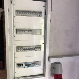 Otwarta skrzynka elektryczna z bezpiecznikami i licznikiem na ścianie, widoczny kabel ochronny i gniazdo siłowe, surowy tynk w tle.