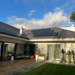 Nowoczesny dom jednorodzinny z panelami słonecznymi na ciemnoszarym dachu, widoczne z drewnianego tarasu z meblami ogrodowymi i dekoracyjnymi trawami na tle błękitnego nieba z chmurami.