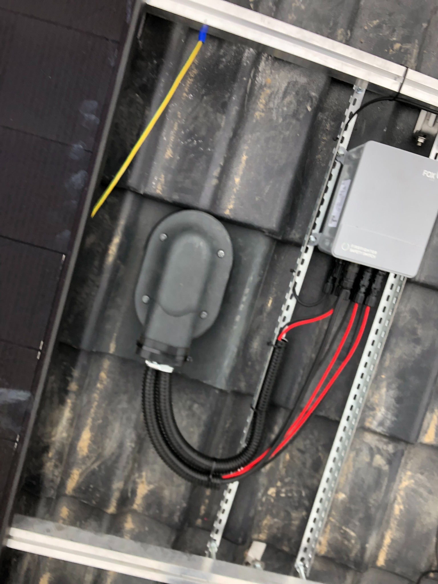 Szara skrzynka elektryczna z podłączonymi czerwonymi i czarnymi kablami zamontowana na metalowej szynie na tle czarnej, starej dachówki ceramicznej. Żółty kabel wystaje zza dachówki.