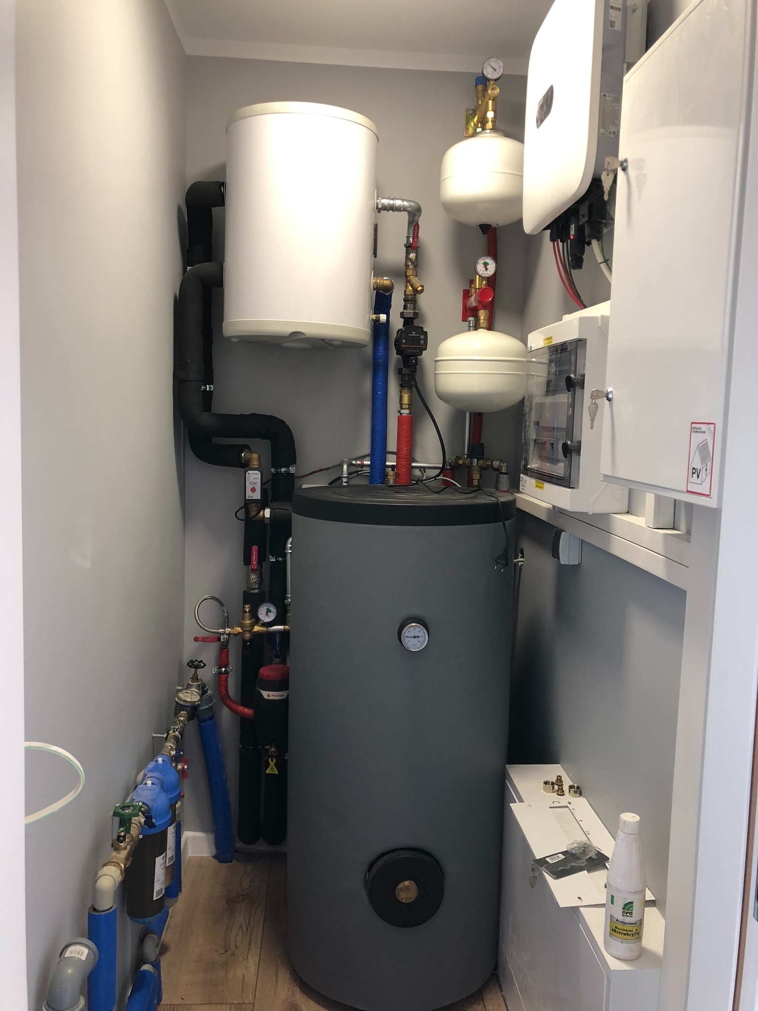 Kompaktowa kotłownia z szarym zasobnikiem ciepłej wody, białym zbiornikiem wyrównawczym i modułem fotowoltaicznym Huawei, z widocznymi rurami, zaworami i manometrami, zainstalowana w małym...