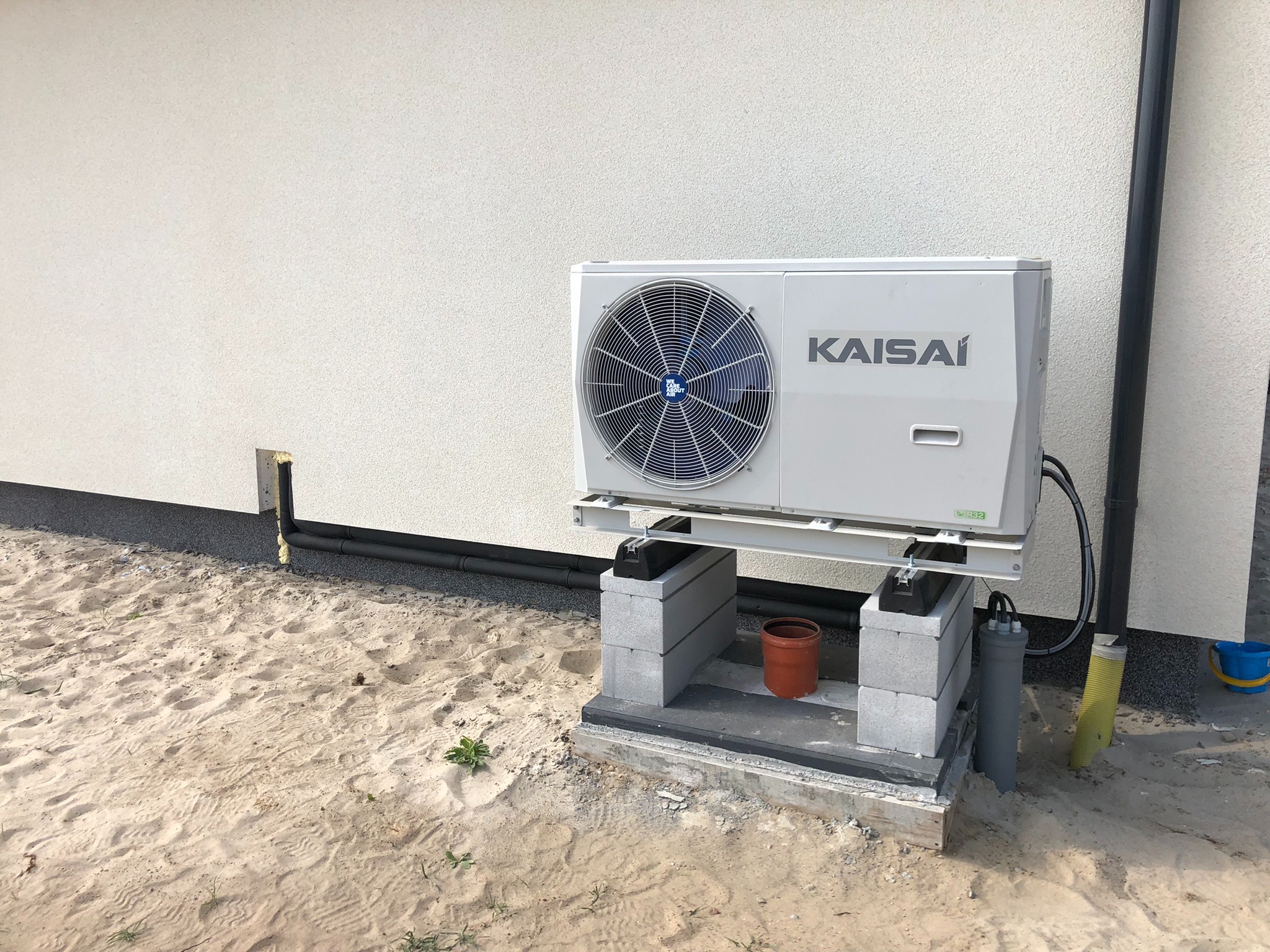 Biała jednostka zewnętrzna z logo Kaisai, zamontowana na betonowych bloczkach na piaszczystym terenie przy ścianie budynku z widoczną rurą odprowadzającą skropliny.