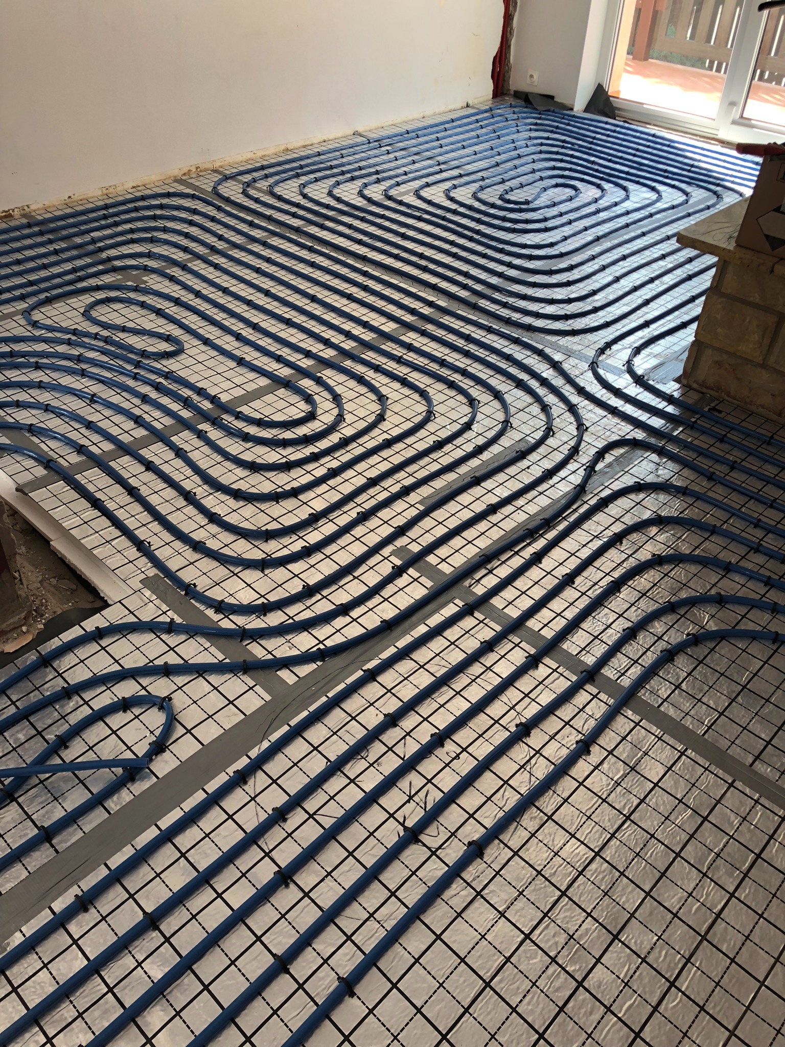 Niebieskie rurki ogrzewania podłogowego ułożone spiralnie na srebrnej folii z czarną siatką, widoczne fragmenty ściany i okna.