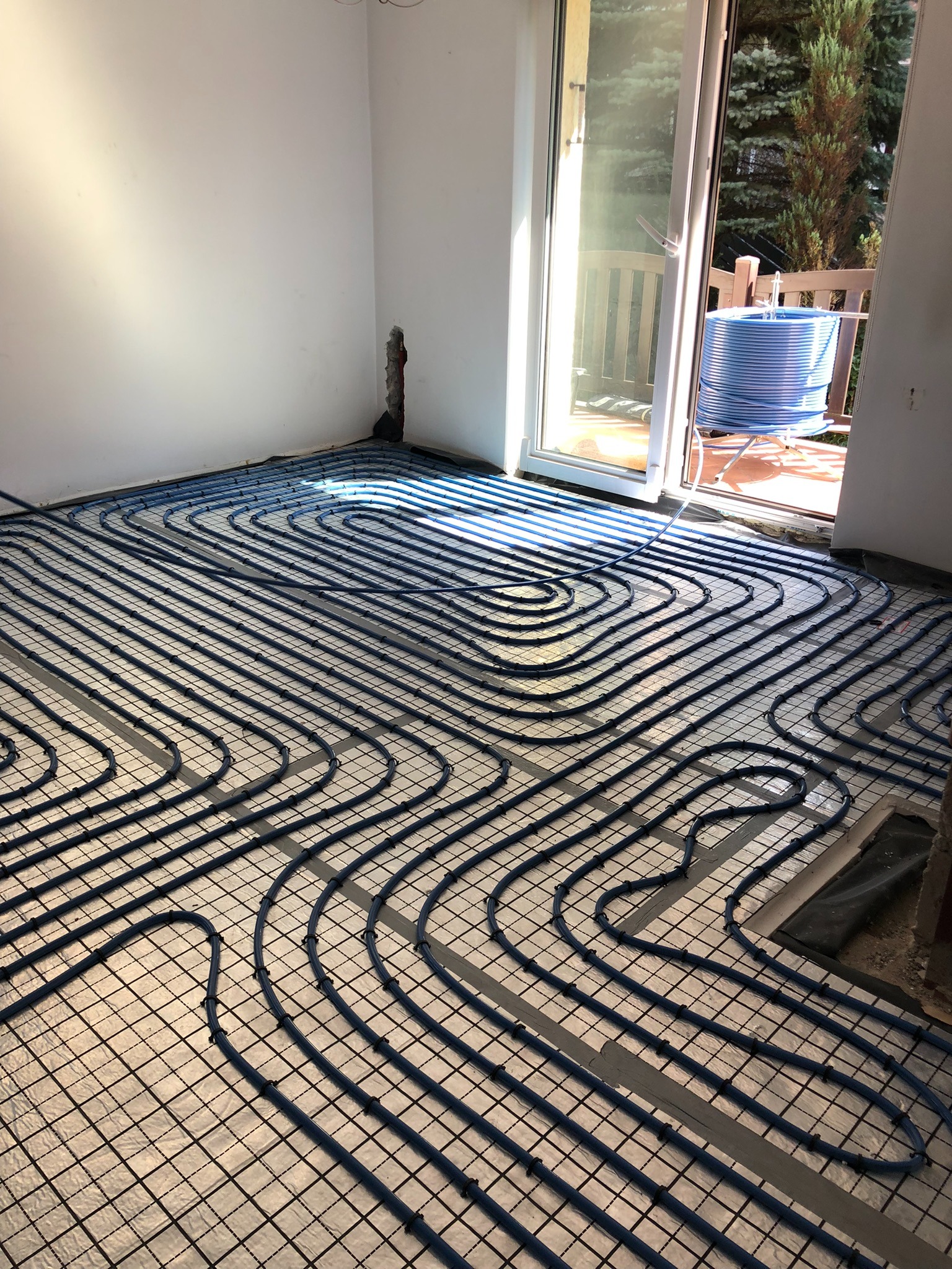 Niebieskie rury ogrzewania podłogowego ułożone na siatce w jasnym pomieszczeniu z otwartym balkonem, w tle zwój rury na balkonie.