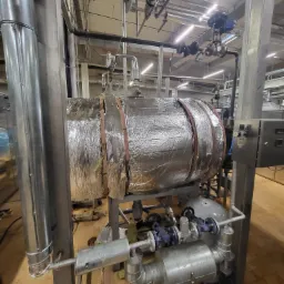 Izolowany zbiornik przemysłowy z instalacją rurową, widoczne zawory i manometry, całość w hali produkcyjnej.