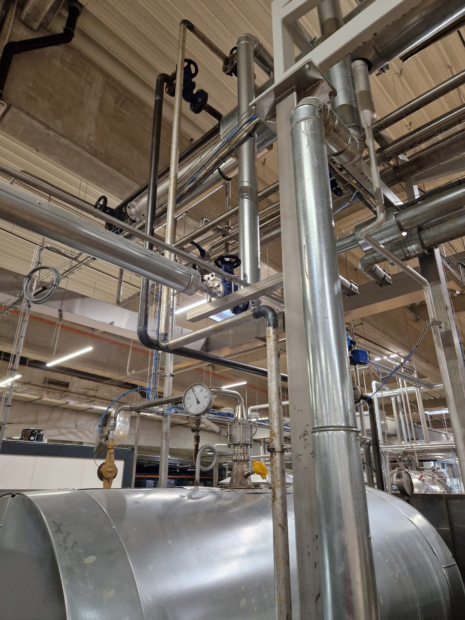 Srebrny zbiornik ciśnieniowy z manometrem i systemem rur w hali przemysłowej, widok z dołu.