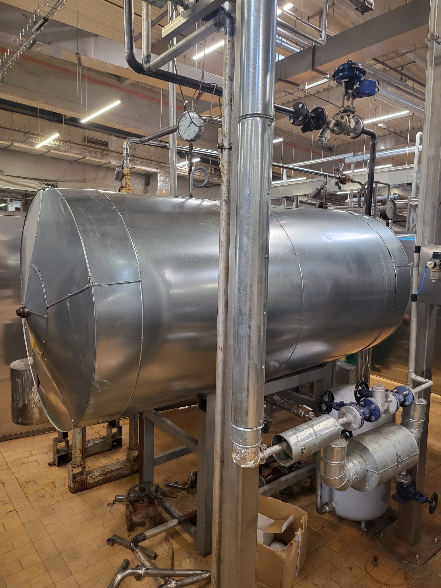 Duży, poziomy, metalowy zbiornik przemysłowy z rurami i zaworami w zakładzie produkcyjnym, widoczne elementy instalacji i podłączeń.