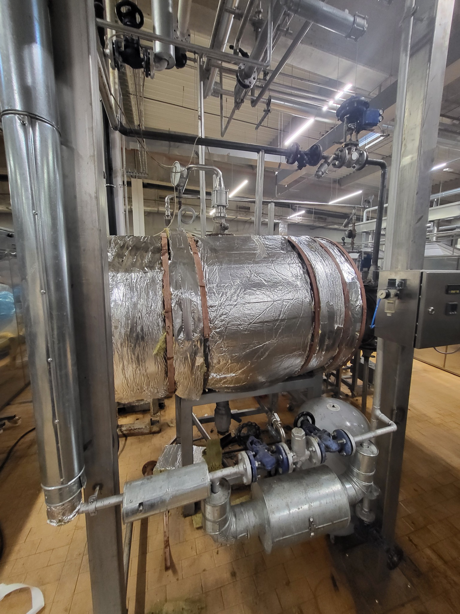 Izolowany zbiornik przemysłowy z instalacją rurową, widoczne zawory i manometry, całość w hali produkcyjnej.