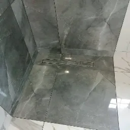 Szara, marmurkowa kabina prysznicowa z odpływem liniowym, wykończona białymi płytkami imitującymi marmur na podłodze poza kabiną. Widoczny fragment buta i rury PCV.