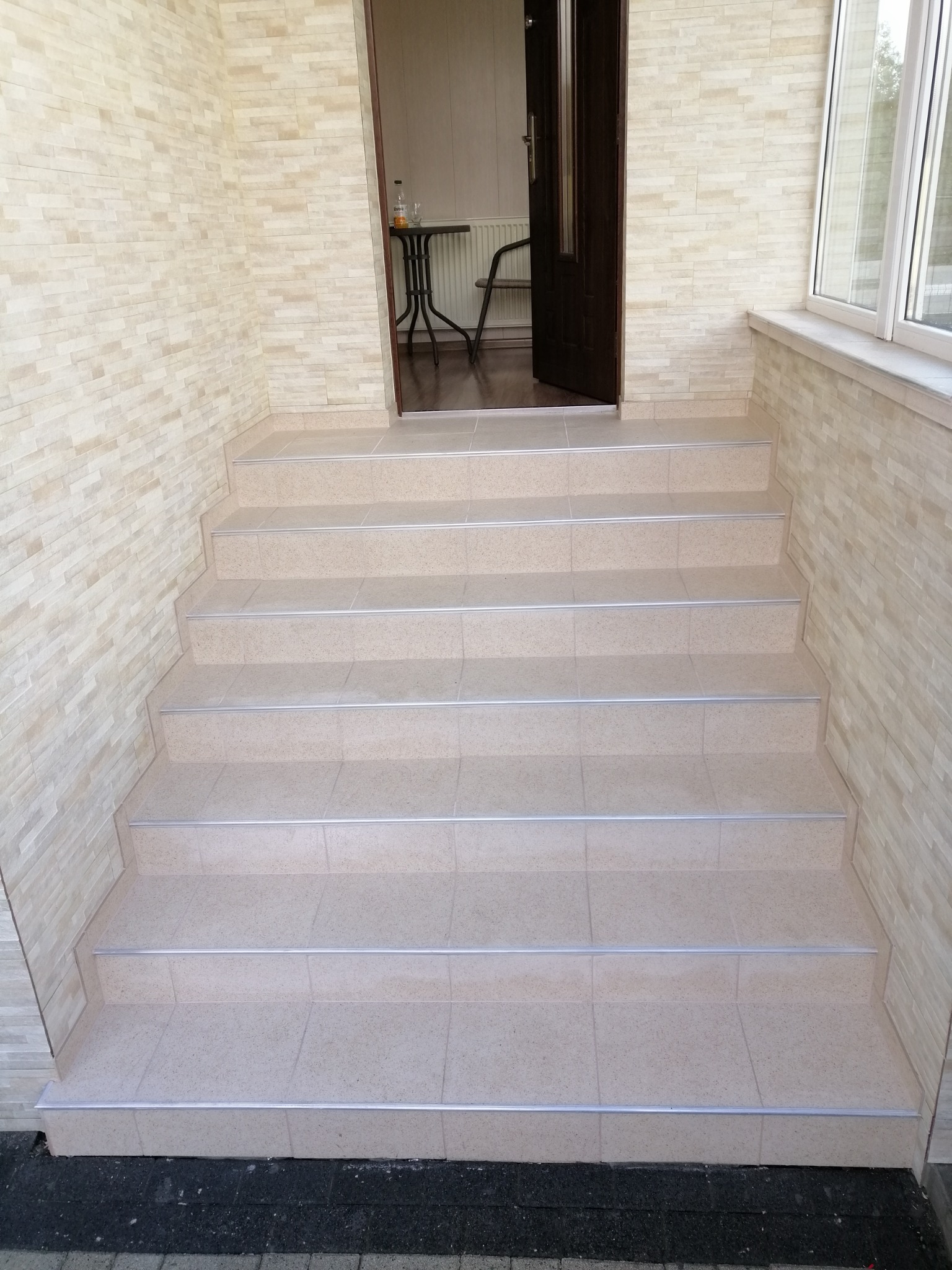 Wykończone płytkami schody wejściowe z metalowymi listwami, ściany wyłożone dekoracyjną cegłą w odcieniach beżu, otwarte drzwi wejściowe.
