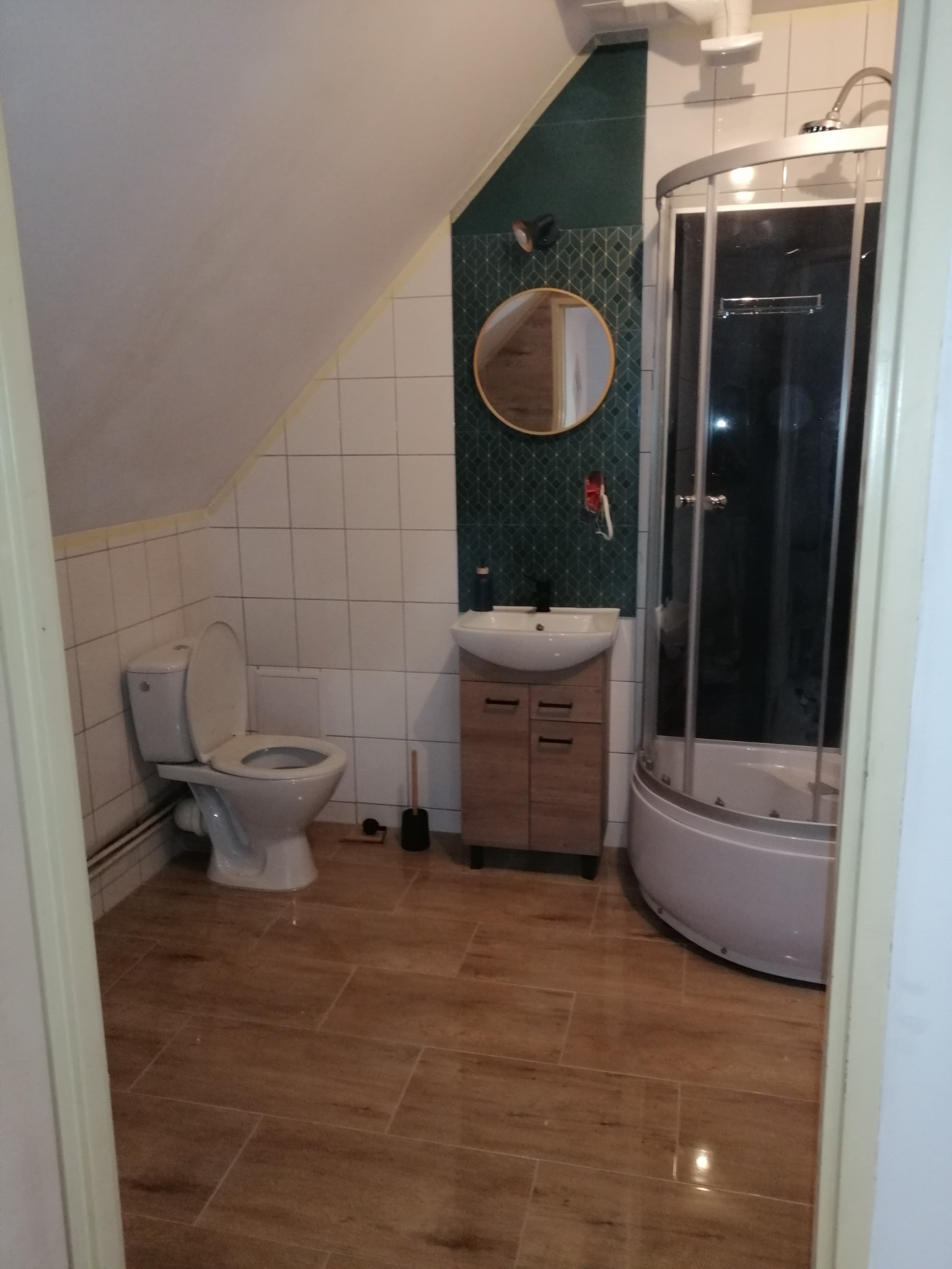 Ukończona łazienka na poddaszu ze skośnym sufitem, z kabiną prysznicową z hydromasażem, umywalką z szafką, okrągłym lustrem, toaletą i brązowymi płytkami imitującymi drewno na podłodze.