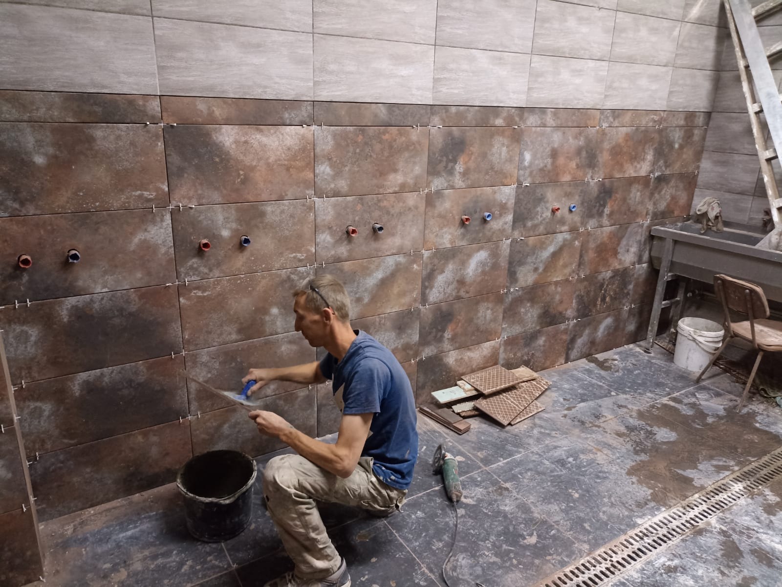 Pracownik w trakcie układania brązowo-szarych płytek na ścianie z wyprowadzonymi instalacjami wodnymi, widoczne krzyżyki dystansowe, obok wiadro z klejem, szlifierka kątowa i drabina