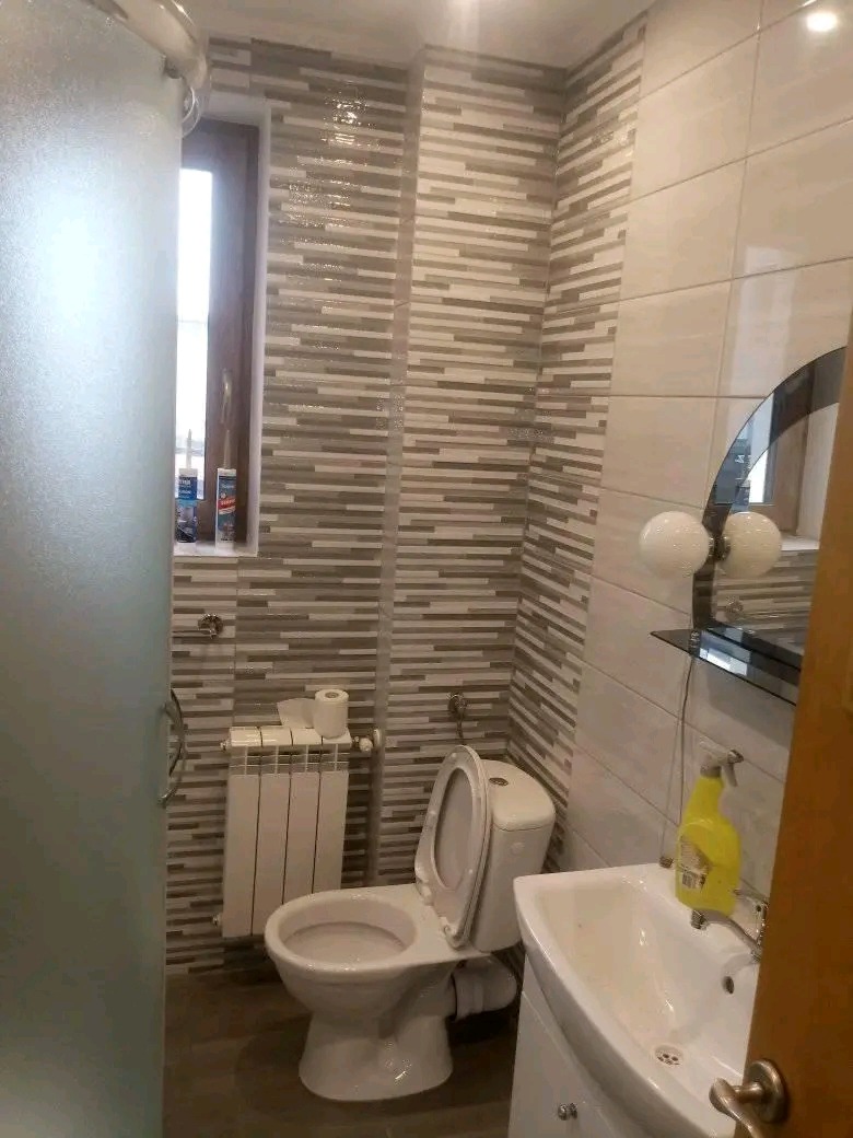 Wykończona łazienka z poziomymi płytkami w odcieniach szarości i beżu, biała toaleta z otwartą klapą, grzejnik żeberkowy, umywalka z szafką i butelka płynu czyszczącego na blacie.