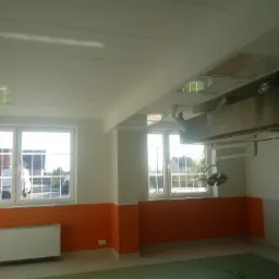 Wnętrze pomieszczenia z oknami i pomarańczową lamperią, widoczne białe płyty sufitowe i metalowa instalacja pod sufitem.