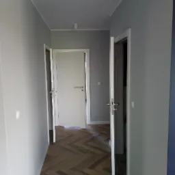 Korytarz z szarymi ścianami, drewnianą podłogą ułożoną w jodełkę i trzema białymi drzwiami z metalowymi klamkami, oświetlony dwoma lampami sufitowymi.