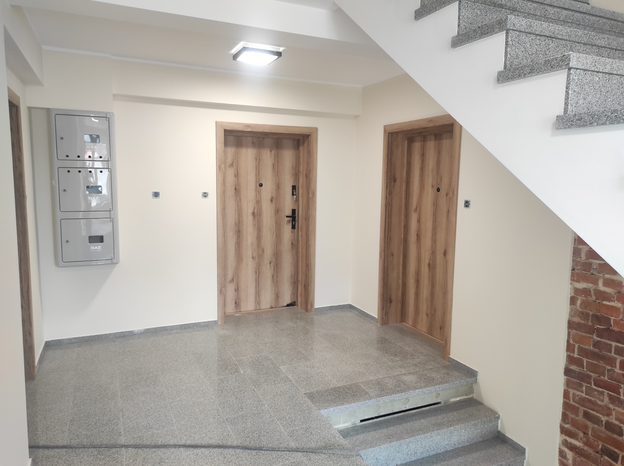 Klatka schodowa w bloku mieszkalnym: szare płytki podłogowe, drewnopodobne drzwi wejściowe, skrzynki gazowe na ścianie, schody z szarego kamienia.