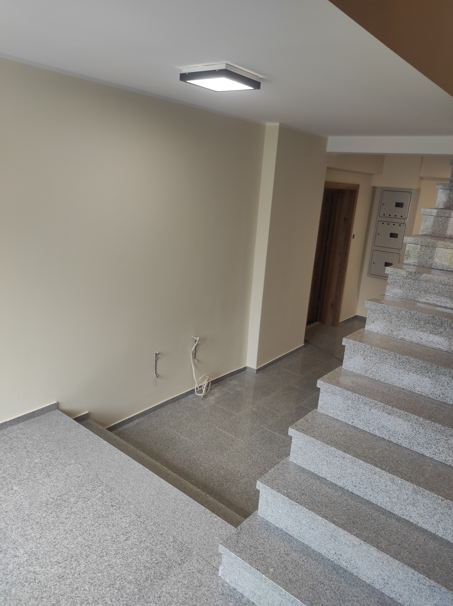 Klatka schodowa w budynku mieszkalnym z wykończonymi ścianami w odcieniach beżu, szare granitowe schody i podłoga, widoczne wystające przewody elektryczne.