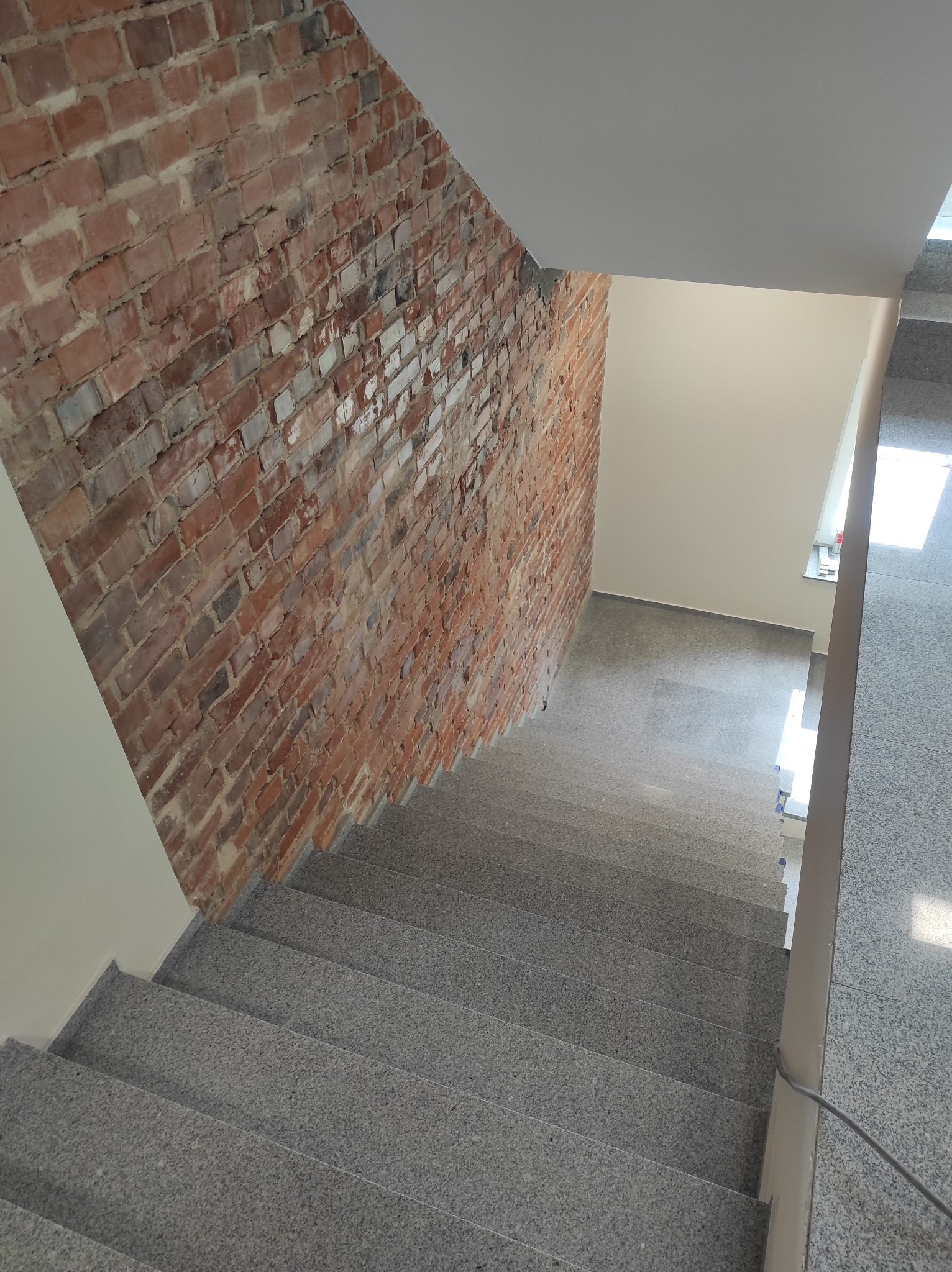 Klatka schodowa z szarymi, granitowymi schodami, ściana z cegły klinkierowej i pomalowana ściana na biało, widok z góry.
