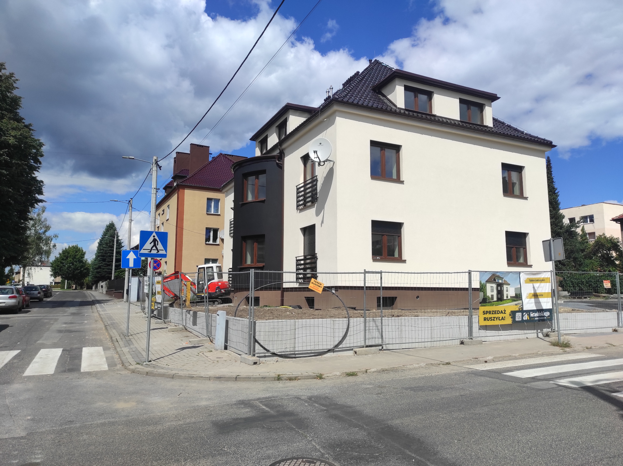 Dwupiętrowy dom z jasną elewacją i ciemnymi oknami, częściowo wykończony, z widocznym mini-ekskawatorem na placu budowy, ogrodzony siatką i tablicą informacyjną.