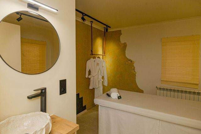 Wnętrze łazienki w spa z okrągłym lustrem, kamienną umywalką, ręcznikiem i dwoma białymi szlafrokami na wieszaku rurowym.