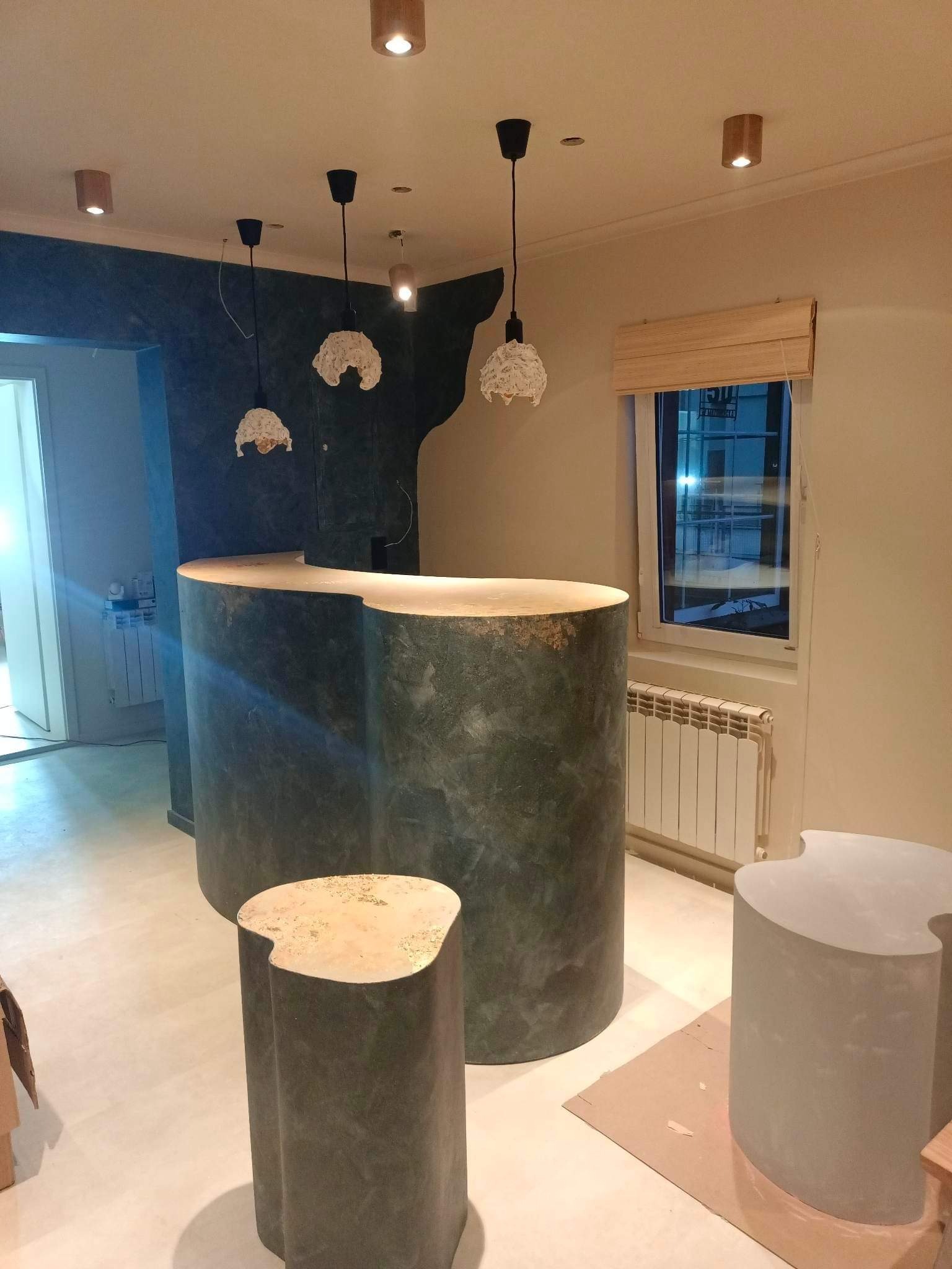 Nowoczesne biuro z zaokrągloną ladą recepcyjną z ciemnego, teksturowanego materiału i designerskimi stołkami w kształcie pni drzew, oświetlone wiszącymi lampami z ażurowymi kloszami.