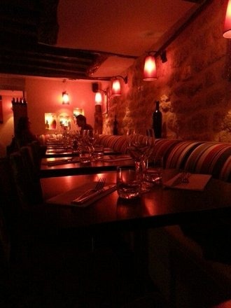 Ustawiony stół w restauracji z ciemnego drewna, ze sztućcami, szklankami i kieliszkami do wina, oświetlony ciepłym, czerwonym światłem, z widocznym fragmentem kamiennej ściany i tapicerowanej ławy...