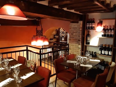 Wnętrze restauracji z czerwonymi akcentami: krzesła, lampy, oraz półki z butelkami wina, widoczne nakryte stoły i elementy dekoracyjne z cegły.