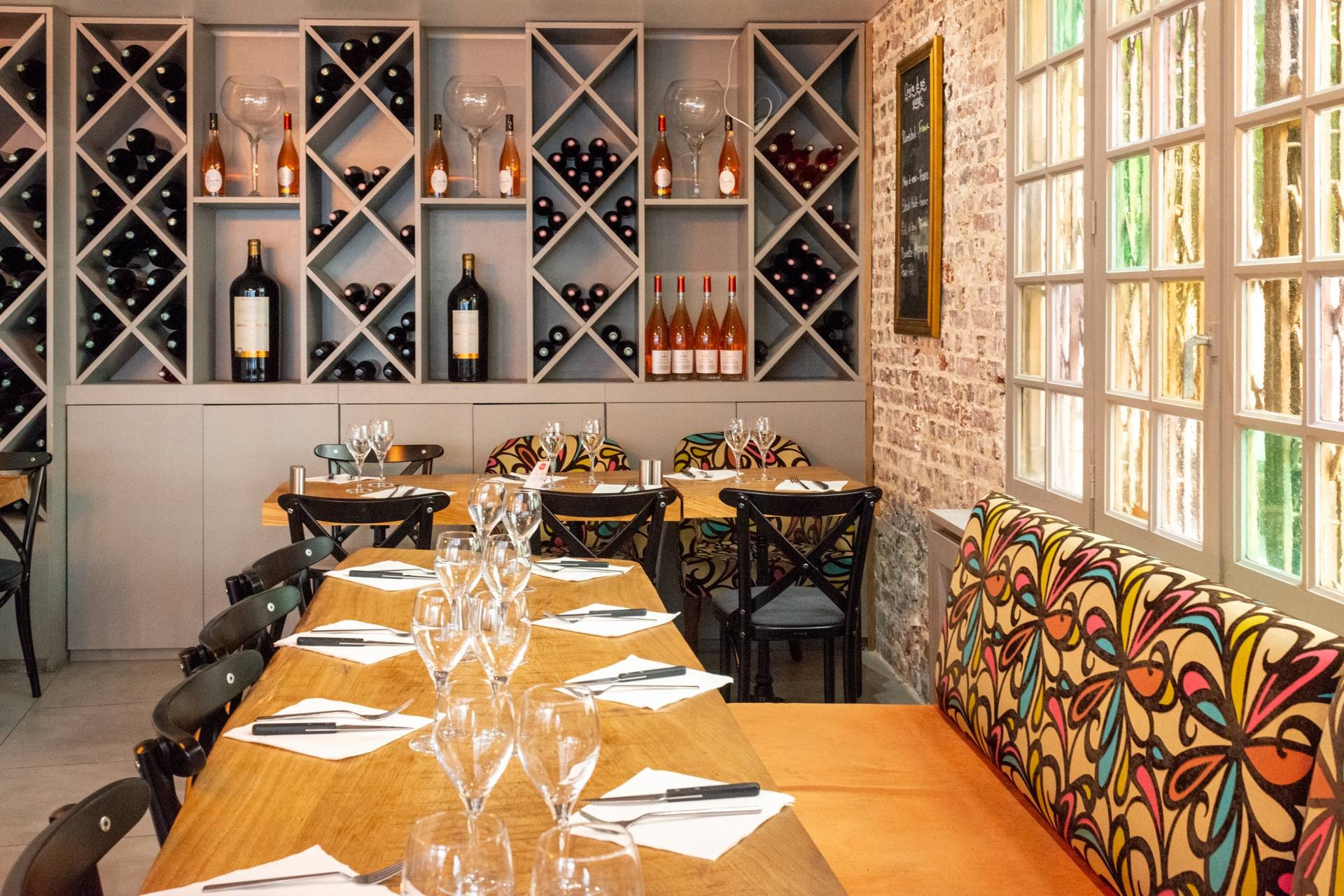 Wnętrze restauracji z ustawionymi stołami, dekoracyjną ścianą z winami i kolorowym siedziskiem pod oknem z witrażami.