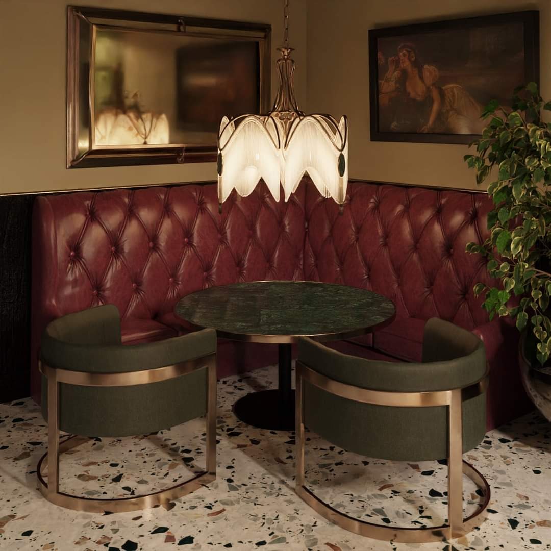 Kącik restauracyjny z pikowaną, bordową kanapą, okrągłym stolikiem z zielonego marmuru i dwoma krzesłami z oliwkową tapicerką i złotymi nóżkami, oświetlony dekoracyjną lampą wiszącą.