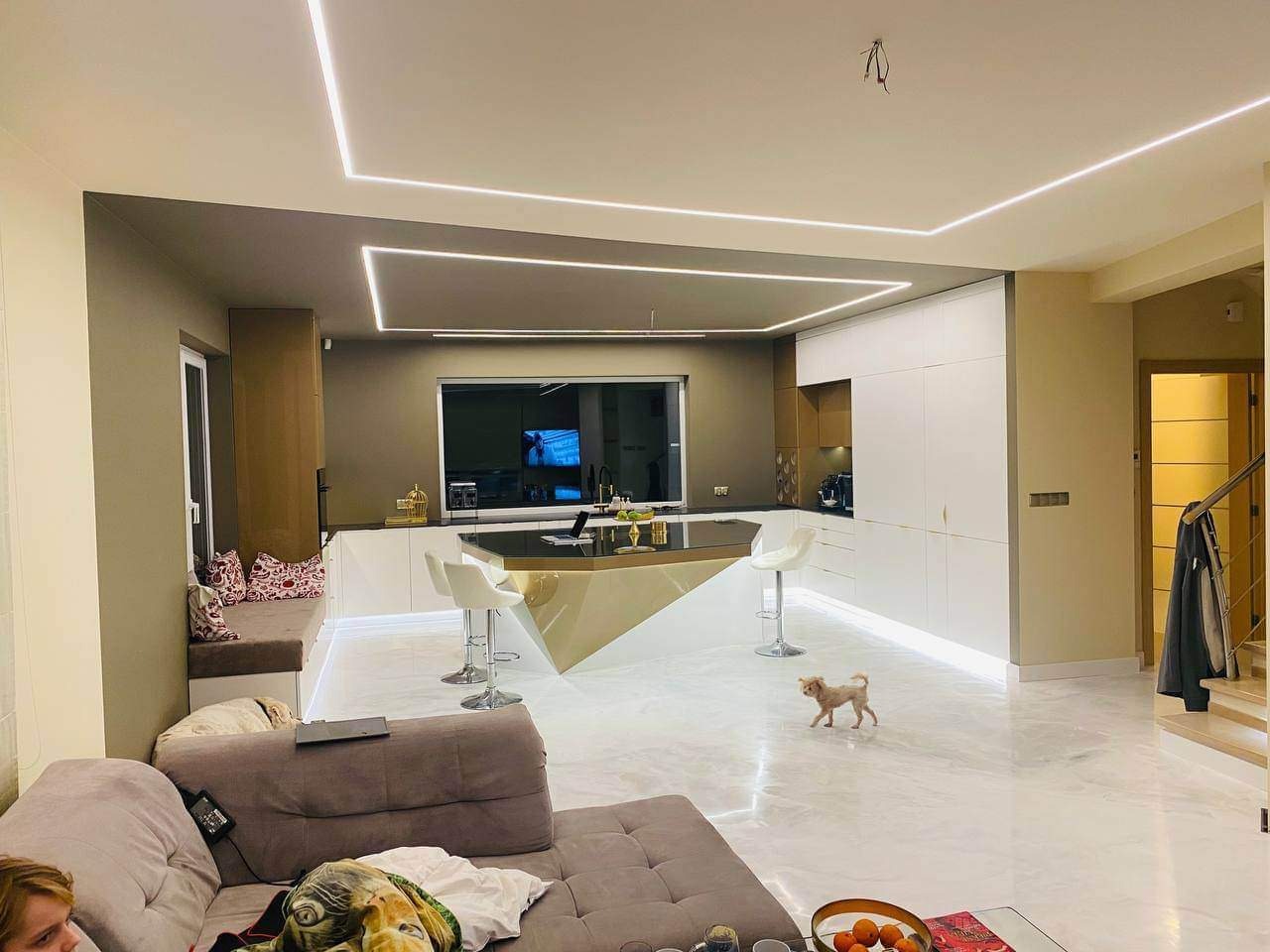Nowoczesne wnętrze salonu z aneksem kuchennym, wyspa kuchenna z czarnym blatem i złotą podstawą, podświetlenie LED sufitu i podłogi, mały pies na marmurowej podłodze, sofa z poduszkami.