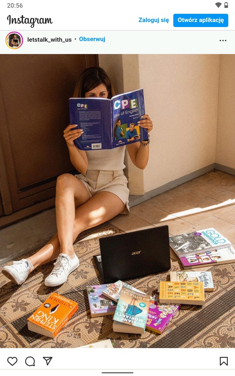 Kobieta siedzi na podłodze, trzyma otwarty podręcznik do angielskiego CPE zasłaniający jej twarz, obok laptop Acer i stos książek, w tym Stephen King 'Outsider' i Liane Moriarty 'Big Little Lies'.