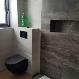 Nowoczesna łazienka z czarną, podwieszaną toaletą, wykończona płytkami imitującymi marmur w odcieniach szarości i bieli, z wnęką w ścianie i butelką wody mineralnej na zabudowie.